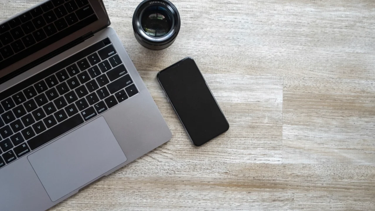 A budget Android smartphone resting on a wooden desk next to a laptop and camera lens