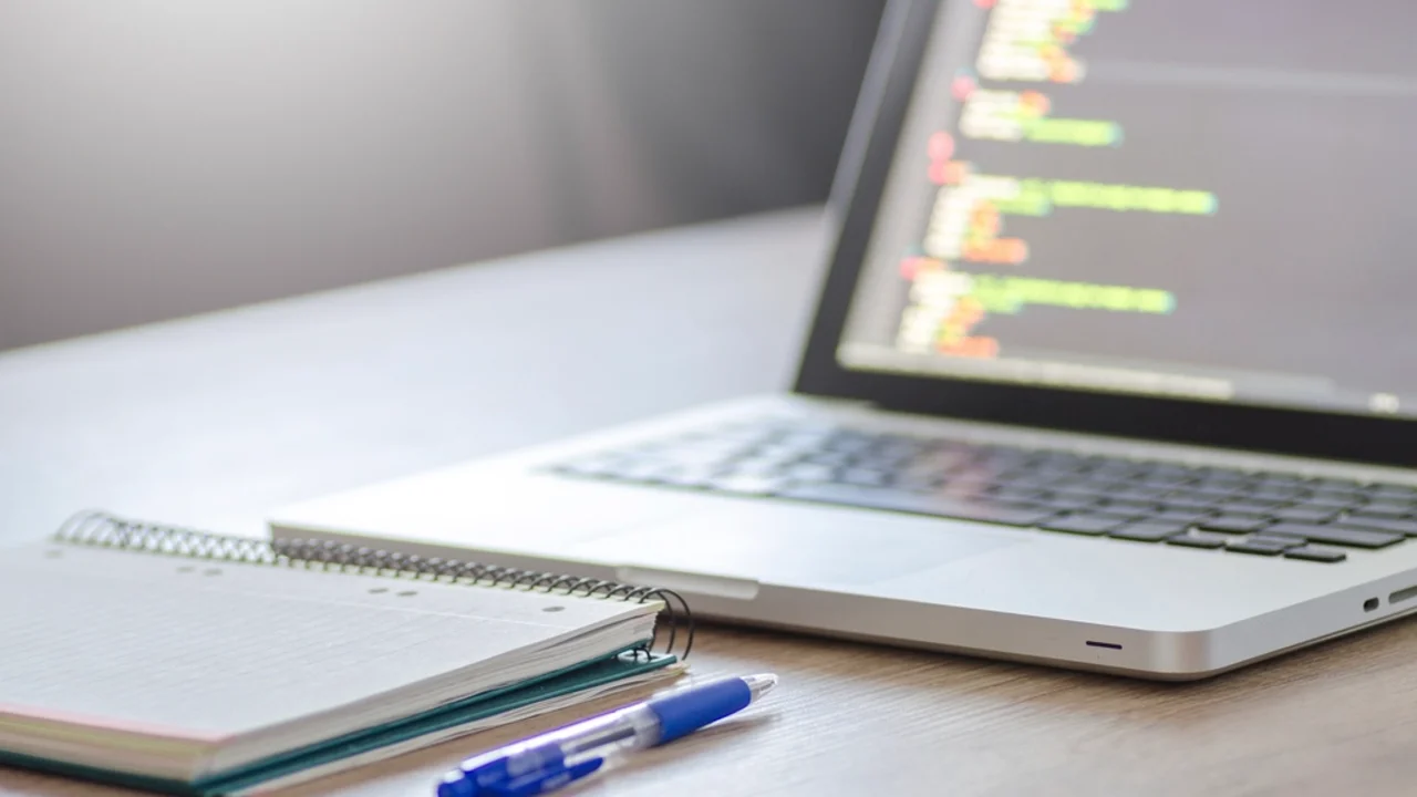 Laptop screen showing colorful code on a wooden desk with a spiral notebook and pens in the foreground