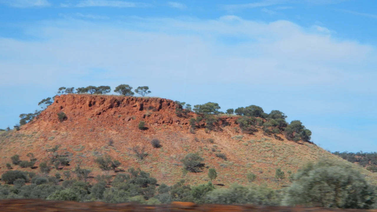 Red rocky outback mesa with sparse green trees and shrubs under a blue sky with light clouds